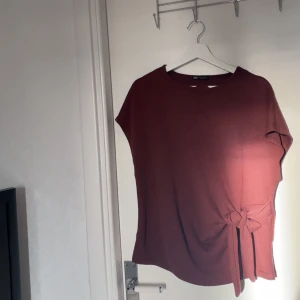 Vinröd topp med knyt från Zara - Säljer en vinröd topp från Zara med rund hals och korta ärmar. Toppen har en snygg knyt-detalj på sidan som ger en cool touch. Materialet känns mjukt och skönt, perfekt för att lyfta din outfit med lite färg.