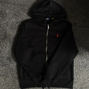 Svart hoodie från Polo Ralph Lauren - Svart hoodie från Polo Ralph Lauren med dragkedja och huva. Klassisk design med röd broderad logga på bröstet och två fickor framtill. Mjuk insida och ribbade muddar. Perfekt för en chill och stilren look.