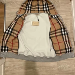 Grå Burberry hoodie med dragkedja - Snygg grå hoodie från Burberry med klassiskt rutigt foder i huvan. Tröjan har dragkedja framtill, snörning i huvan och ribbade muddar. Perfekt för dig som vill ha en stilren men ändå lyxig look. Ikoniskt Burberry-mönster på insidan av huvan. Vi har många storlek 