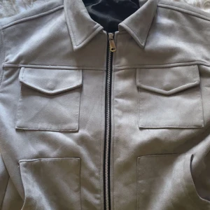 Beige mocka overshirt med dragkedja - Snygg beige overshirt i mockaimitation med klassisk krage, två stora bröstfickor med lock och dragkedja framtill. Perfekt lager-på-lager-plagg med stilren och minimalistisk look.