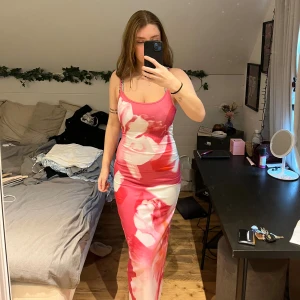 Rosa blommig långklänning med slits - Säljer en snygg långklänning med blommigt mönster i rosa, vitt och rött. Klänningen har smala axelband, figurnära passform och en slits på sidan. Materialet känns mjukt och stretchigt.