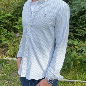 Randig Ralph Lauren Skjorta - Säljer en stilren och snygg blåvit randig skjorta från Ralph Lauren. Storlek S (custom fit), men sitter mer som en M slim fit. Skjortan är i mycket fint skick och passar utmärkt, både till vardags och mer uppklädda tillfällen.  Har du några frågor? Tveka inte att höra av dig!Allt gott från Deluxe-garderoben