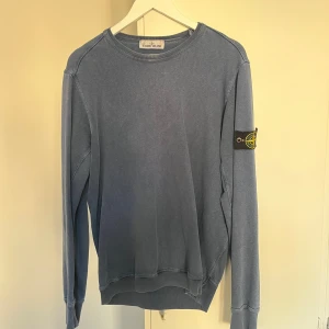 Blå sweatshirt från Stone Island - Säljer en blå sweatshirt från Stone Island med klassisk patch på vänster ärm. Tröjan har rund hals, långa ärmar och ribbade muddar. Tillverkad i mjuk bomull och har en relaxed passform som är perfekt för en chill stil.