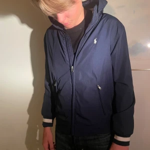  Windbreaker Polo Ralph Lauren - Snygg mörkblå vindjacka från Polo Ralph Lauren med huva, dragkedja framtill och två fickor med dragkedja. Jackan har ribbade muddar med vit rand och klassisk logga på bröstet. Perfekt för blåsiga dagar och enkel att matcha med det mesta.