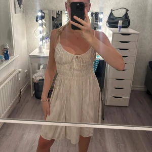 ljusbeige/vit Randig kort klänning från Nelly - Supersöt kort klänning med smala axelband och ljusa beige-vita ränder. Klänningen har snörning framtill vid bysten och smockad midja som ger en skön passform. Materialet är mjukt och lätt, perfekt för varma dagar. Enkel och fräsch look som är lätt att styla. Knappt använd 🤍🤎