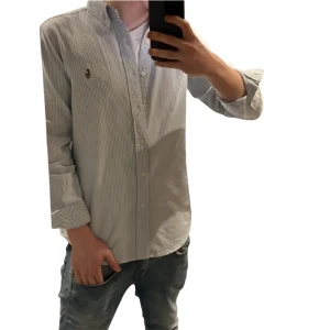 Randig skjorta från Polo Ralph Lauren - Klassisk skjorta från Polo Ralph Lauren i vitt och blått med smala ränder. Skjortan har lång ärm, knappar framtill och broderad logga på bröstet. Tillverkad i mjuk bomull och har en normal passform med button down-krage.