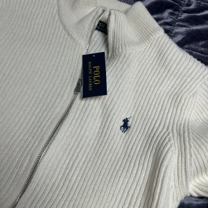 Vit stickad kofta från Polo Ralph Lauren - Vit ribbstickad kofta från Polo Ralph Lauren med dragkedja framtill och klassisk broderad logga på bröstet. Tröjan har lång ärm och hög krage, perfekt för lager-på-lager. Snygg och stilren design som passar till allt.