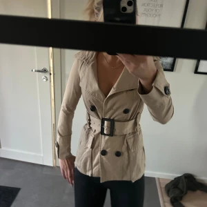 Beige trenchcoat från Zara Woman - Snygg beige trenchcoat från Zara Woman med klassisk dubbelknäppning, stor krage och bälte i midjan med stor spänne. Jackan har långa ärmar, fickor med lock och svarta knappar som kontrast. Ny utan prislapp.
