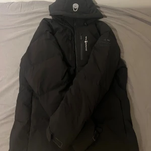Svart dunjacka Sail Racing L - Svart dunjacka från Sail Racing, modell Antarctica Expedition. Jackan har huva, flera dragkedjor och coola logodetaljer på bröstet och ärmen. Fylld med Allied Feather + Down, 500 fill power. Perfekt för kalla vinterdagar och har en clean, sportig look. Bara använd 3 gånger.