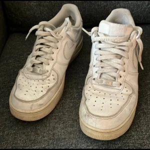 Nike Air Force 1 vita sneakers i storlek 44 - Klassiska Nike Air Force 1 sneakers i helvitt skinn med perforerad tå och platt sula. Snygg låg modell med snörning och ikonisk Nike-logga på sidan och hälen. Perfekt för dig som gillar streetstyle och vill ha en tidlös sneaker i garderoben.