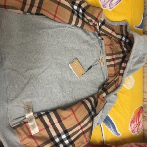 Grå Burberry hoodie med klassiskt rutmönster - Grå hoodie från Burberry med ikoniskt rutigt foder i huvan och insidan av ärmarna. Tröjan har broderad logga på bröstet, huva och klassisk passform. Tillverkad i mjuk bomull och polyesterblandning. Perfekt för dig som vill ha en stilren och lyxig streetlook och den är helt ny oanvänd 