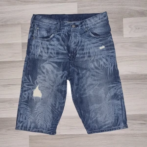 Blå denimshorts med bladmönster H&M - Snygga blå denimshorts från H&M med coolt bladmönster i ljusare toner. Klassisk femficksmodell med knapp och dragkedja fram. Materialet är bomull och shortsen har en midwaist passform. Perfekt för dig som gillar streetstyle och vill sticka ut lite extra.