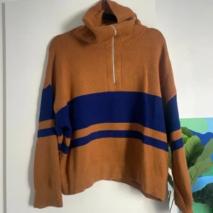 Brun och blå stickad tröja från MNG - Säljer en brun stickad tröja från mango med breda blå ränder och half zip-dragkedja vid halsen. Tröjan har hög krage, lång ärm och en loose passform. 