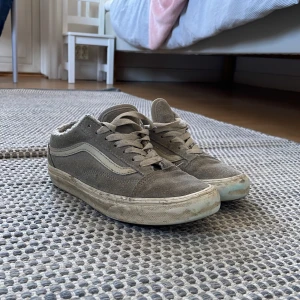 Grå Vans Old Skool sneakers - Säljer ett par grå Vans Old Skool sneakers med klassisk vit sidorand och brun gummisula. Skorna har snörning och är tillverkade i mocka och textil. Perfekt för dig som gillar streetstyle och skate-vibe. Låg modell med rund tå och platt sula.