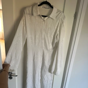  långklänning  - Säljer en elegant vit långklänning från NA-KD med subtilt mönster i tyget. Klänningen har klassisk skjortkrage, knappar hela vägen framtill och långa ärmar med lätt puff. Materialet är mjukt och har en satinliknande finish som ger en lyxig känsla. Går ner till anklarna. En knapp saknas syns på bild två men går enkelt att sy på med knappen som tillkommer se bild 3.
