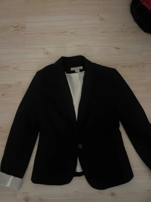 Svart kavaj från H&M, strl 34 - Klassisk svart kavaj från H&M i storlek 34. Enkelknäppt med en knapp framtill och ljus innerfoder som syns när ärmarna viks upp. Snygg passform och tidlös look, perfekt till skjorta eller t-shirt. Kort i armarna!!
