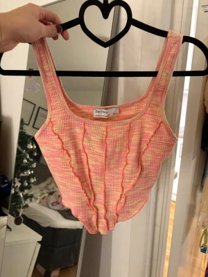 Rutig ribbad linnetopp från Bershka - Säljer en söt rutig linnetopp från Bershka i rosa, gul och orange toner. Toppen har ribbad struktur, breda axelband och dekorativa vågiga sömmar framtill. Perfekt för sommardagar och ger en färgglad vibe till din outfit.