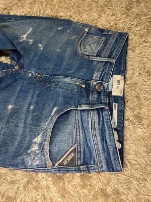 Replay Anbass blå jeans - Säljer ett par Replay Anbass jeans i klassisk blå tvätt med slitningar och detaljerade sömmar. Jeansen har normal passform, fem fickor och Replay-logga på fickan. Perfekta för dig som gillar en avslappnad men snygg stil. Det är slim fit