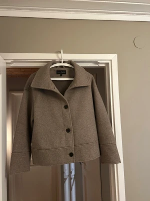 Beige jacka från KappAhl i S - Stilren beige  jacka från KappAhl i storlek S. Jackan har en rak, boxig passform med stora knappar framtill och bred krage. Tillverkad i en mjuk blandning av polyester, akryl och viskos. Perfekt för lager-på-lager.