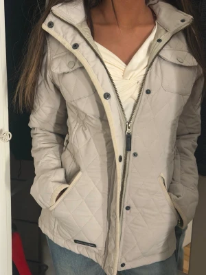 Beige quiltad jacka från Trespass - Snygg beige quiltad jacka från Trespass med huva och fuskpälsdetalj. Jackan har dragkedja, knappar, två fickor och justerbart bälte i midjan, och sitter tajt runt midjan. Perfekt för kyligare dagar och har en stilren look med rutmönster och långa ärmar.