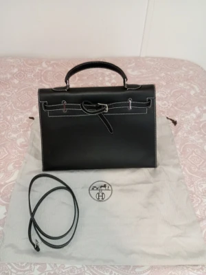 Svart handväska vintage 2007 - Säljer en svart handväska i slätt skinn med vit kontrastsöm och silverfärgade metalldetaljer. Väskan har ett klassiskt handtag, avtagbar axelrem och elegant spänne framtill. Insidan är rymlig och stilren, perfekt för dig som gillar lyxiga accessoarer.