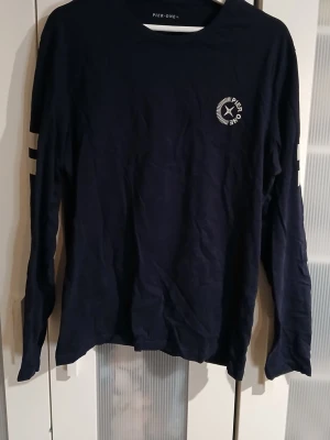 Mörkblå långärmad topp Pier One - Mörkblå långärmad topp från Pier One med vitt tryck på bröstet och två vita ränder på ena ärmen. Klassisk rund halsringning och rak passform. Tillverkad i mjuk bomull, perfekt för en chill och stilren look. Känns mer som en medium!