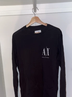 Svart långärmad t-shirt Armani Exchange - Svart långärmad t-shirt från Armani Exchange med vit logga på bröstet. Klassisk rund halsringning och stilren design. Perfekt för dig som gillar snygga och enkla plagg med premiumkänsla. Materialet är mjukt och skönt, troligtvis bomull.