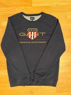 Svart sweatshirt från GANT - Säljer en svart sweatshirt från GANT med broderad logga och emblem i guld, rött och vitt på bröstet. Tröjan har rund hals, ribbade muddar och lång ärm. Perfekt för dig som gillar klassisk amerikansk stil och vill ha något snyggt och bekvämt.