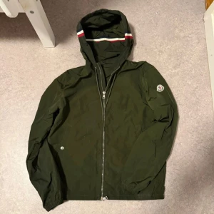 Moncler windbreaker - Snygg mörkgrön vindjacka från Moncler med huva och dragkedja framtill. Jackan har Moncler-logga på ärmen och coola ränder i rött, vitt och blått på huvan. Perfekt för dig som gillar stilrena och sportiga plagg med exklusiv känsla. Storlek nummer 1, skick 9/10, för mer bilder kom priv.