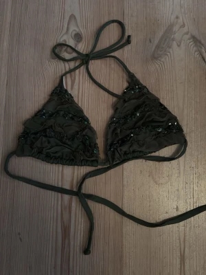 Grön paljett bikiniöverdel Bluepoint - Säljer en grön bikiniöverdel från Bluepoint med volangdetaljer och paljetter. Triangelformad modell med knyt i nacke och rygg. Perfekt för dig som vill ha något extra på stranden.