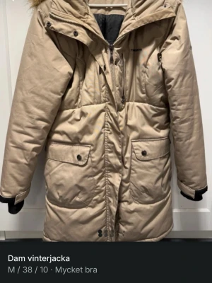 Beige vinterparkas från Trionarth - Säljer en beige vinterparkas från Trionarth med huva och fuskpäls. Jackan har flera fickor med både dragkedja och knappar, samt ribbade muddar vid ärmslut. Perfekt för kalla dagar och har en snygg, lång passform.