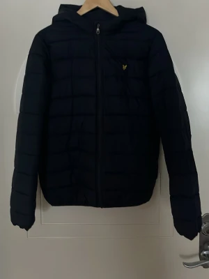 Svart pufferjacka med huva från Lyle & Scott - Säljer en mörknlå pufferjacka från Lyle & Scott med huva, dragkedja framtill och två sidofickor. Jackan har det klassiska guldiga logomärket på bröstet. Den är lite sönder på magen och är sydd på ena armen men detta är inget som syns tydligt. Priset kan diskuteras. Står storlek 14-15 år  men är som en xs