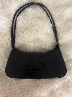 Svart axelväska med rosettdetalj - Snygg svart axelväska i skinnimitation med elegant rosettdetalj framtill. Väskan har dubbla axelband, magnetlås och innerfack med dragkedja. Perfekt storlek för det viktigaste och stilren design som passar till många outfits.