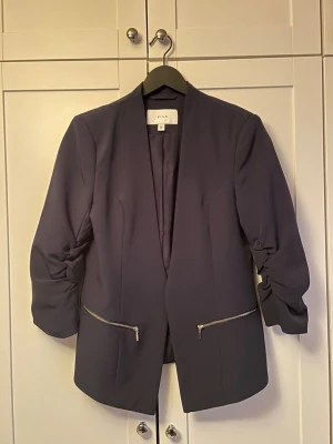 Marinblå kavaj från Vila med dragkedjor - Säljer en marinblå kavaj från Vila med snygga dragkedjor framtill och rynkade ärmar. Kavajen har en stilren, öppen design utan krage och är tillverkad i ett mjukt syntetmaterial. Perfekt för dig som vill ha en clean look med edge.