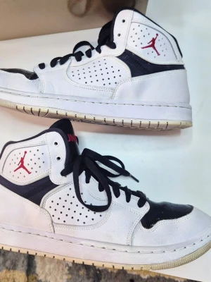 Nike Air Jordan 1 High vit/svart - Säljer ett par Nike Air Jordan 1 High i vitt och svart skinn med röda detaljer och svart snörning. Klassisk perforerad tåbox, högt skaft och Jumpman-logga på plösen och sidan. Perfekta för dig som gillar streetstyle och vill sticka ut.