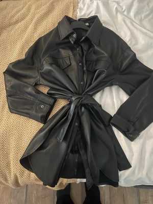 Svart skinn overshirt med knyt - Snygg svart overshirt i skinnimitation med knyt i midjan och klassisk krage. Jackan har långa ärmar, tryckknappar framtill och två bröstfickor med lock. Perfekt för att ge din outfit en edgy vibe.