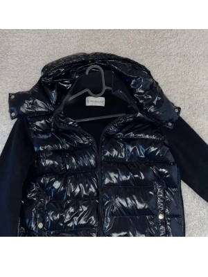 Moncler pufferjacka - Säljer en mörkblå pufferjacka från Moncler med glansig yta och quiltad design. Jackan har avtagbar huva, dragkedja framtill och ribbade muddar. Perfekt för kyliga dagar och riktigt snygg till streetwear-stilen. De enda ”felet” är att den har ett litet märke på framsidan (se bild 3). Syns inte vid användning förutom om man kollar riktigt noga. Pris är alltid diskuterbart🙌🏽