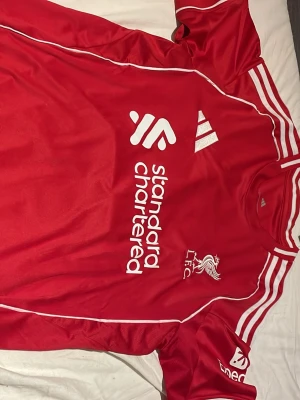Liverpool tröja Adidas M Salah 11 - Liverpool FC matchtröja från Adidas i röd med vita detaljer och tryck. Slim fit-modell med M. Salah och nummer 11 på ryggen. Officiell produkt med Standard Chartered sponsortryck och Aeroready-material för extra komfort.