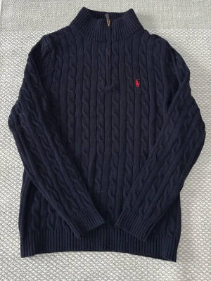 Marinblå stickad tröja Polo Ralph Lauren - Kabelstickad marinblå tröja från Polo Ralph Lauren med halv dragkedja och ribbad hög krage. Klassisk röd broderad logga på bröstet. Tillverkad i mjuk bomull och har långa ärmar samt ribbade muddar för extra komfort.