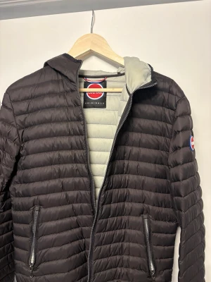 Svart pufferjacka från Colmar - Säljer en svart pufferjacka från Colmar med huva och två dragkedjeförsedda fickor. Jackan har quiltad design, Colmar-logga på ärmen och ljusgrått innerfoder. Perfekt för kyliga dagar och riktigt snygg till vardagslooken.