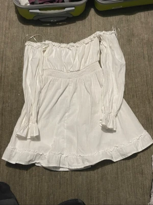 Vit offshoulder klänning från Amisu - Supersöt vit kortklänning från Amisu med offshoulder och långa ärmar. Klänningen har smock i midjan, volangkant nertill och är tillverkad i mjuk bomull. Perfekt för sommarens alla tillfällen!