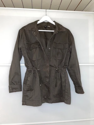 Mörkgrön Parkajacka - Säljer en parkajacka från H&M Divided i storlek S. Jackan har dragsko i midjan, fyra fickor framtill och knappar vid ärmsluten. Perfekt för lager-på-lager och snygg till våren/hösten✨😊