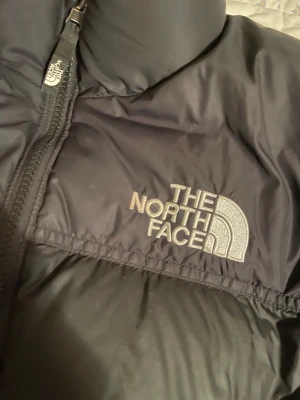 Svart pufferjacka från The North Face XL - Säljer en svart pufferjacka från The North Face i storlek XL för ungdom. Jackan har hög krage, två sidofickor med dragkedja och broderad logga på bröstet. Finns ett litet ihopsytt hål på ena armen. Materialet är polyester och jackan har en klassisk quiltad look med hel dragkedja framtill.