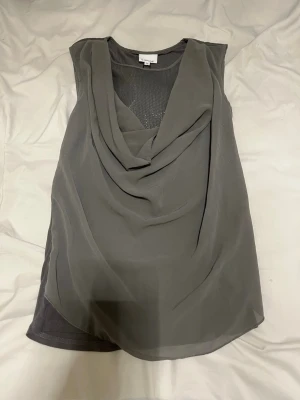 Grå ärmlös blus från b.young - Säljer en snygg grå ärmlös blus från b.young i modellen Georgette. Toppen har ett draperat fall framtill och är gjord i ett tunt, lätt material som ger en luftig känsla. Perfekt till både jeans och kjol för en stilren look.