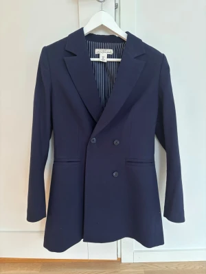 Marinblå kavaj från L.O.G.G. strl 38 - Snygg marinblå kavaj från L.O.G.G. i storlek 38. Dubbelknäppt framtill med krage och två fickor. Insidan har ett randigt foder i vitt och blått. Perfekt för dig som gillar liiiite längre och mjukare kavajer!