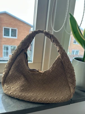 Beige flätad axelväska - Säljer en snygg beige axelväska med flätad design och rundad form. Väskan har ett kort handtag och stängs med dragkedja. Perfekt för dig som vill ha en trendig och stilren accessoar till din outfit.