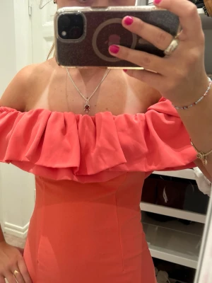 Korallfärgad offshoulder klänning - Säljer en snygg korallfärgad klänning med offshoulder-modell och bred volang runt axlarna. Klänningen är figurnära och har en stilren, enkel design utan mönster. Perfekt för dig som vill sticka ut med färg och volangdetalj.