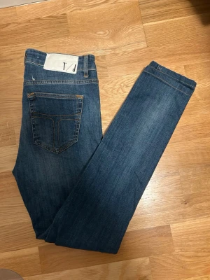 Blå jeans från Tiger of Sweden - 29/34 - Snygga blå jeans från Tiger of Sweden med klassisk femficksdesign och orangea sömmar. Jeansen har rak passform och normal midja, tillverkade i slitstarkt bomullsmaterial. Perfekta för dig som gillar stilrena och tidlösa jeans.