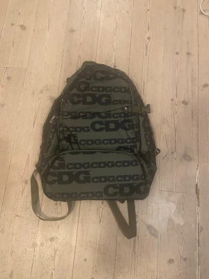 CDG ryggsäck (Comme des Garçons)  - Säljer en snygg ryggsäck från Comme des Garçons  med stort svart logomönster över hela väskan. Utsidan är olivgrön med svarta detaljer och insidan är knallorange. Väskan har flera fack, justerbara remmar och dragkedjor. Perfekt för dig som gillar streetwear och vill sticka ut.