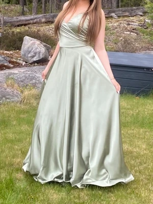 Ljusgrön långklänning i satin - Elegant ljusgrön långklänning i glansigt satinmaterial med v-ringad omlottdesign upptill och axelbandslös skärning. Kjolen är vid och faller snyggt mot marken, perfekt för bal eller student. Nypris 3300 köpt på ivory Field Stockholm. Använd en gång och kemtvättad efter användning så helt ren och bra skick